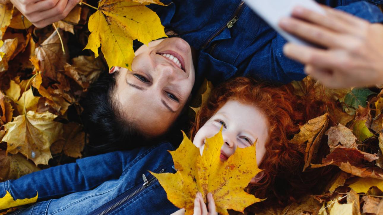 A woman and her daughter lie on the ground surrounded by colorful autumn leaves, enjoying a moment together