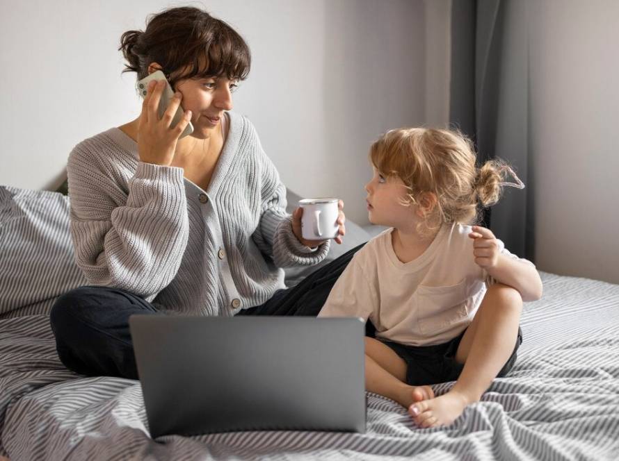 A woman and a child are seated on a bed, engaged with a laptop and sipping coffee in a relaxed atmosphere
