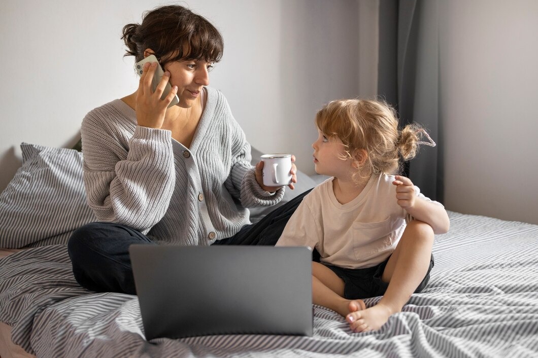 A woman and a child are seated on a bed, engaged with a laptop and sipping coffee in a relaxed atmosphere